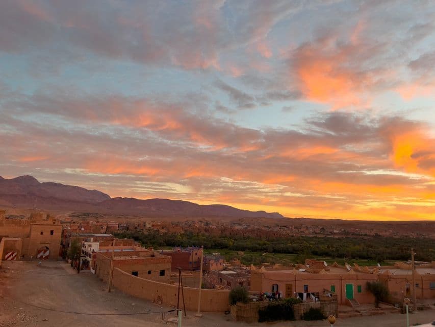 Un villaggio di edifici in terra si trova in una valle ai piedi delle montagne, sotto un cielo con nuvole arancioni e rosa al tramonto.