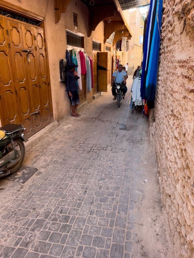 Un uomo in moto percorre un vicolo stretto e acciottolato, fiancheggiato da negozi che espongono abiti e tessuti in vendita.