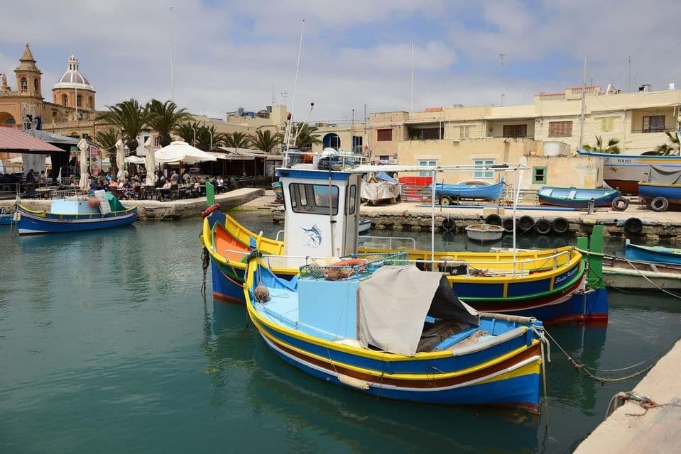 Barcos de pesca coloridos amarrados en un puerto, con edificios frente al mar y una iglesia con cúpula visibles al fondo.