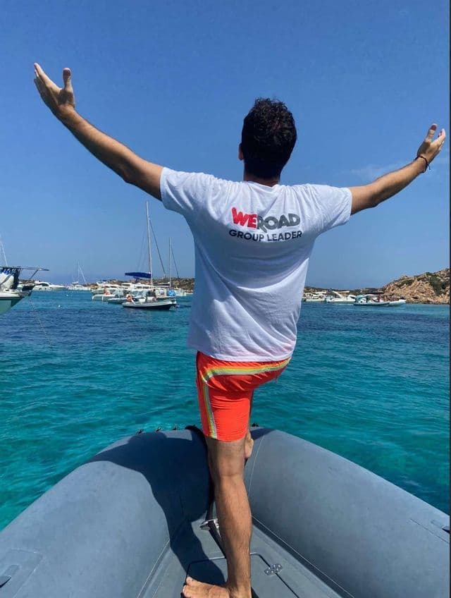 Un líder de grupo WeRoad de pie con los brazos extendidos en la proa de una lancha inflable en agua turquesa.