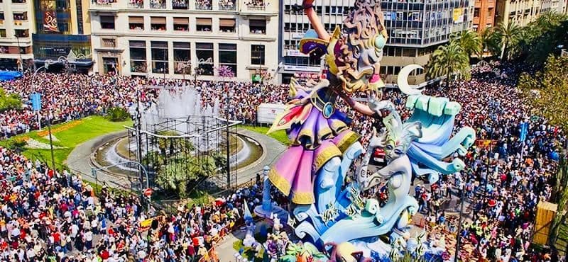 Una folla immensa riempie una piazza cittadina, circondando un grande e elaborato carro allegorico e una fontana centrale in una giornata di sole.