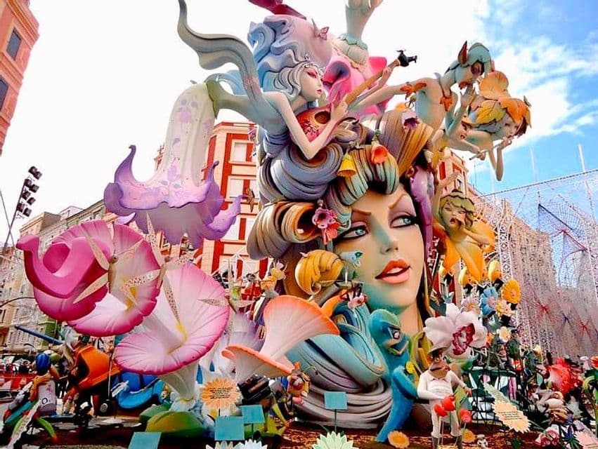 Una scultura imponente e colorata, raffigurante un grande volto femminile, figure fantastiche e fiori, si erge all'aperto in una città.