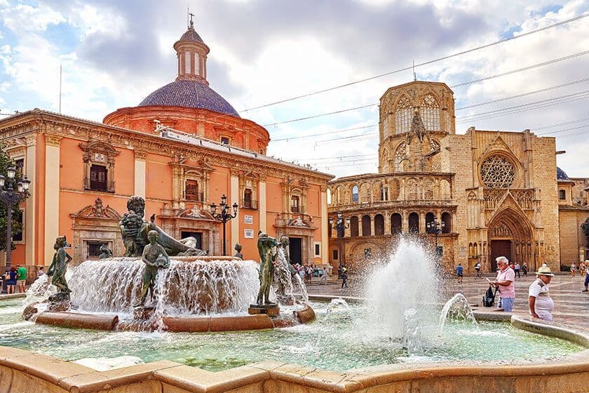 Una grande fontana con statue di bronzo zampilla in una piazza storica, con una cattedrale e un edificio a cupola sullo sfondo.