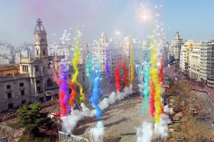Fumi color arcobaleno eruttano da fuochi d'artificio su una piazza affollata durante un festival diurno.