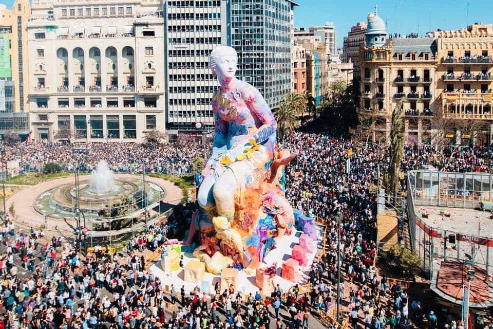Una grande statua colorata di una figura seduta in una piazza cittadina, circondata da una folla immensa durante un festival soleggiato.