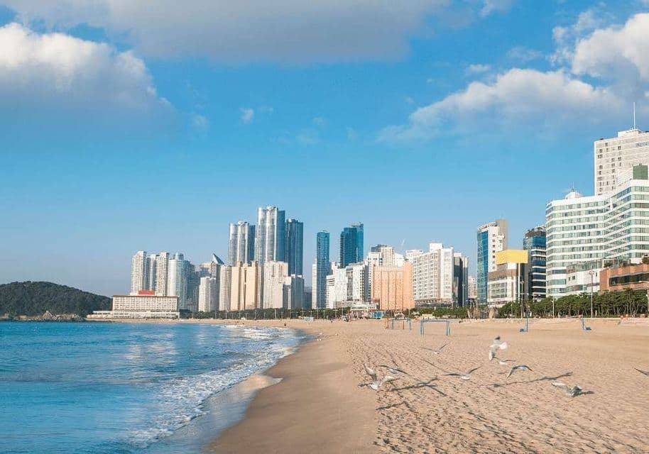 Un'ampia spiaggia sabbiosa con onde dolci incontra uno skyline urbano moderno sotto un cielo blu con nuvole sparse.