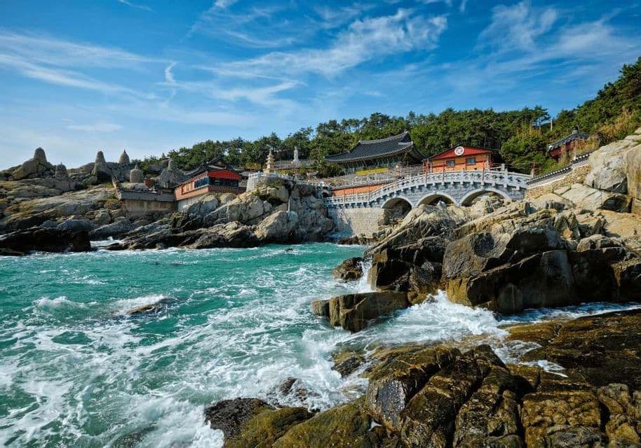 Un tradizionale complesso di templi e un ponte di pietra ad arco si ergono su una costa rocciosa sopra un mare turchese agitato.