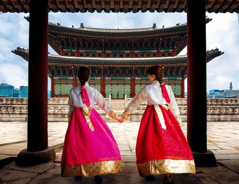 Due donne di spalle, in colorati abiti tradizionali hanbok, si tengono per mano e guardano un grande palazzo.