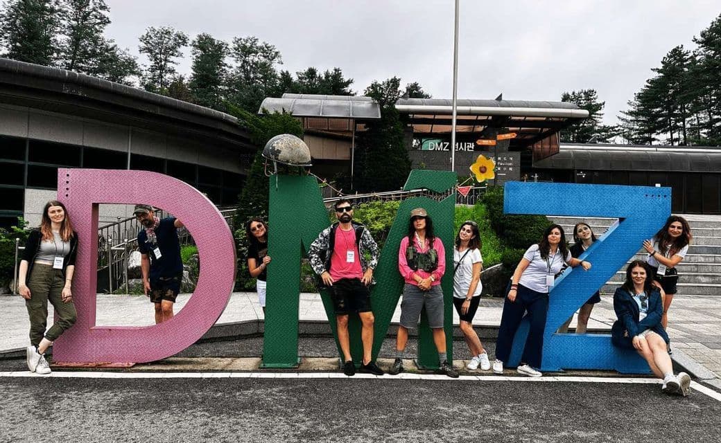 Un viaggio di gruppo WeRoad in posa per una foto con grandi lettere colorate che compongono la scritta DMZ, con un edificio sullo sfondo.