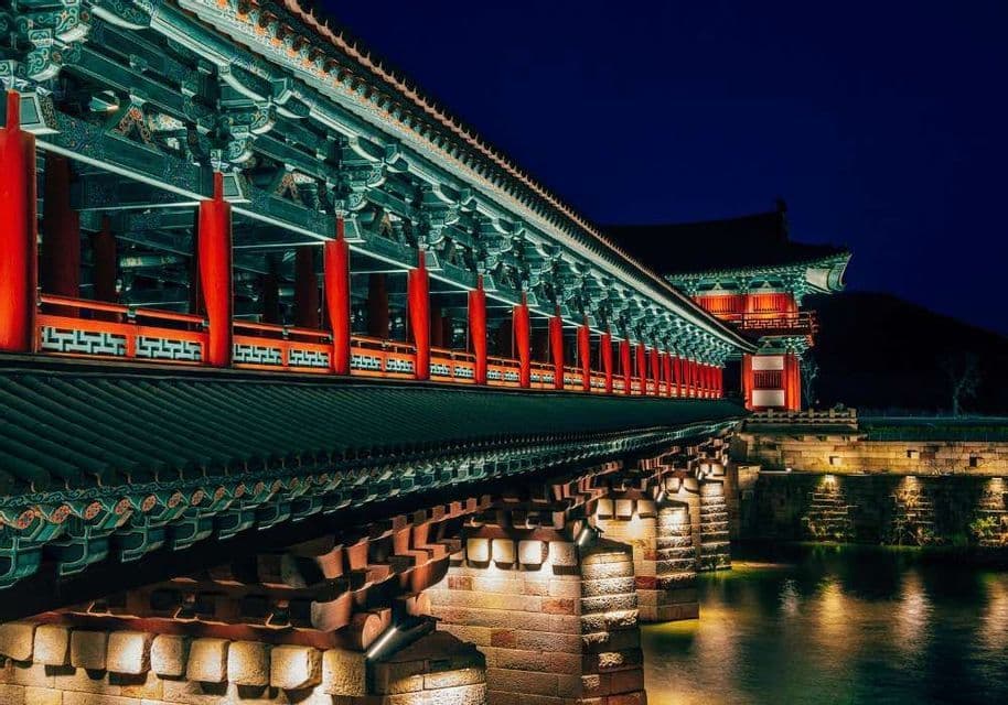 Un ponte tradizionale in stile asiatico con pilastri rossi e tetto di tegole verdi, illuminato di notte sull'acqua