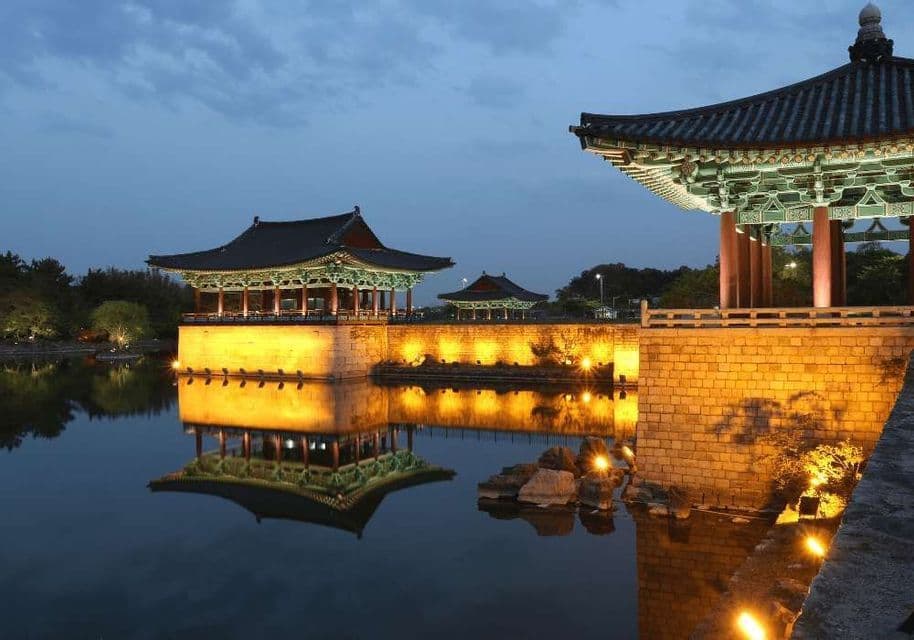 Padiglioni asiatici tradizionali su fondamenta in pietra sono illuminati al crepuscolo, riflettendosi nell'acqua calma sottostante.