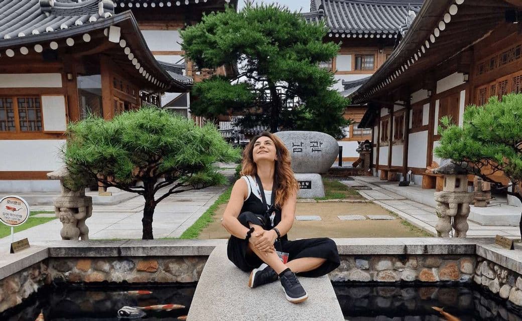 Una donna siede su un cornicione di pietra sopra un laghetto koi, guardando in alto in un cortile tradizionale coreano con alberi bonsai.