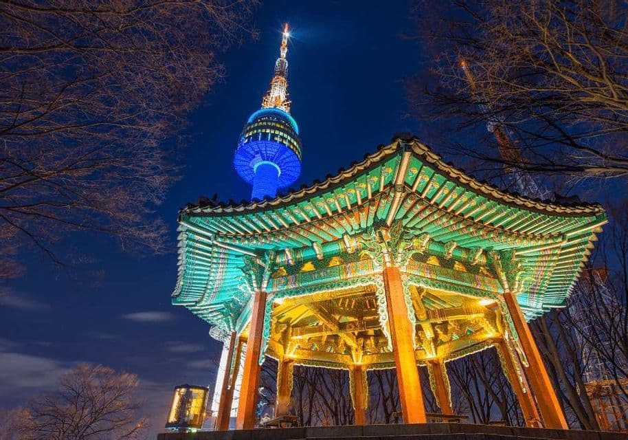 Un padiglione tradizionale dal tetto verde illuminato di notte, con un'alta torre moderna illuminata di blu sullo sfondo sotto un cielo scuro.