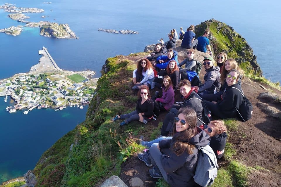 Eine WeRoad-Reisegruppe sitzt lächelnd auf einem grünen Berggipfel mit Blick auf ein Küstendorf und das Meer.
