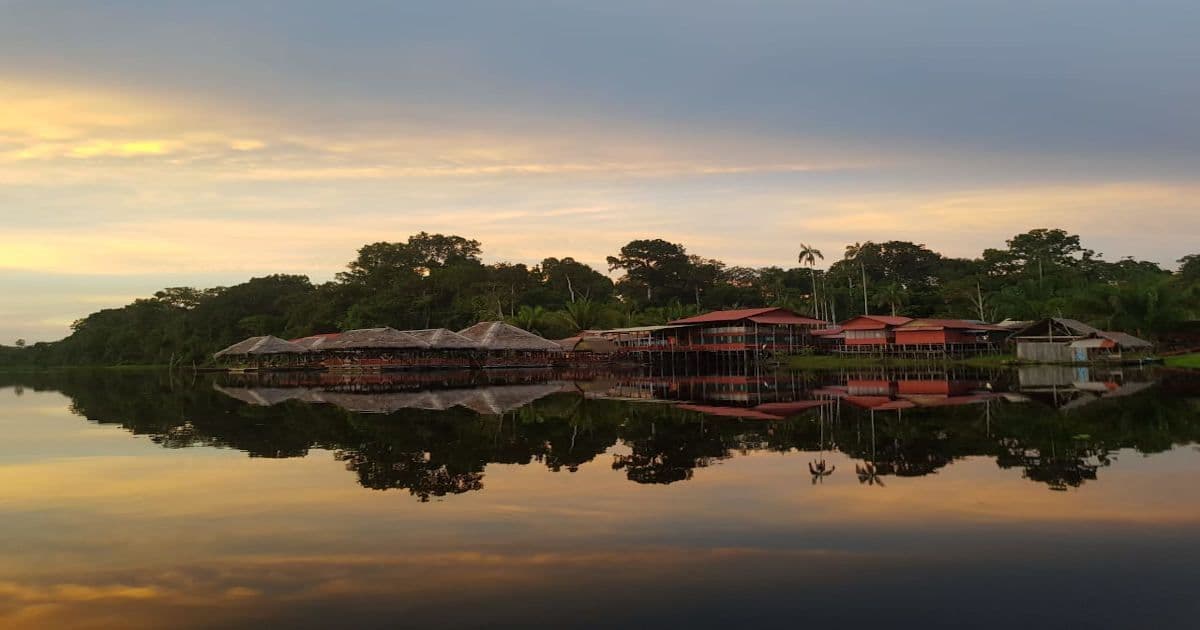 Casette in legno su palafitte vicino a una fitta foresta, perfettamente riflesse nelle acque calme di un lago al tramonto.
