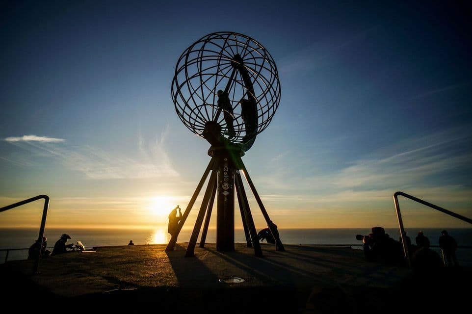 Un voyage de groupe WeRoad en silhouette devant un coucher de soleil, près d'une grande sculpture de globe dominant l'océan.
