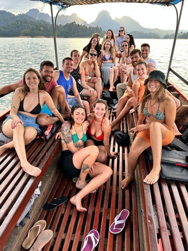 Un grupo de WeRoad en un largo barco de madera, sonriendo a la cámara con un lago y montañas de fondo.