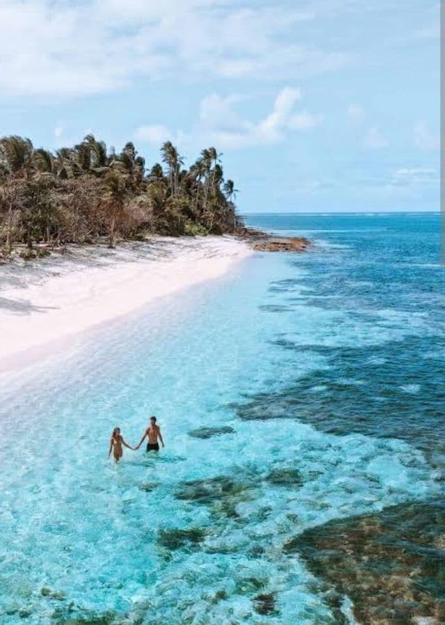 Vista aérea de una pareja cogida de la mano de pie en aguas poco profundas, claras y turquesas frente a una playa de arena blanca con palmeras.
