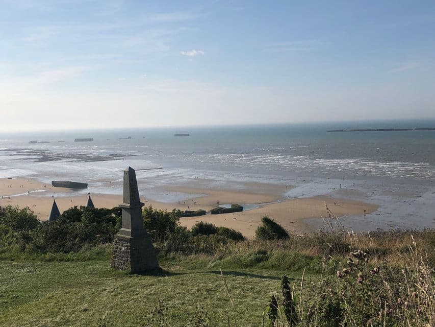Ein Steinmonument auf einem grasbewachsenen Hügel überblickt einen Strand mit Betonstrukturen und das Meer bei Ebbe.