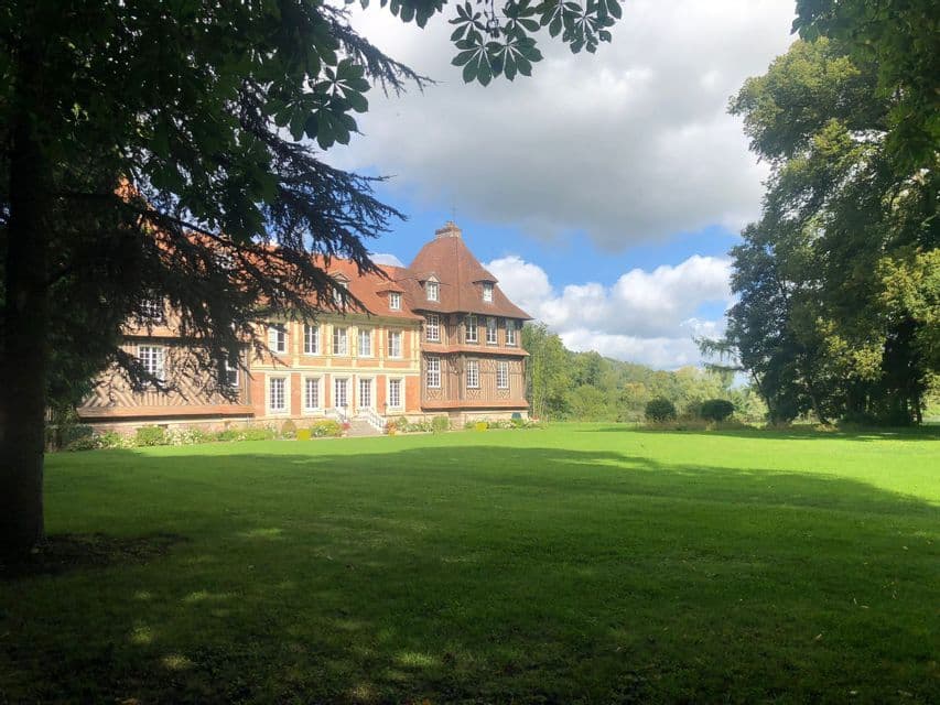 Ein großes Fachwerk-Herrenhaus mit rotem Ziegeldach steht neben einem weiten grünen Rasen, umrahmt von belaubten Ästen.
