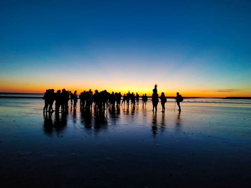 Un groupe WeRoad en silhouette au coucher du soleil sur une plage humide, avec leurs reflets sur le sable.