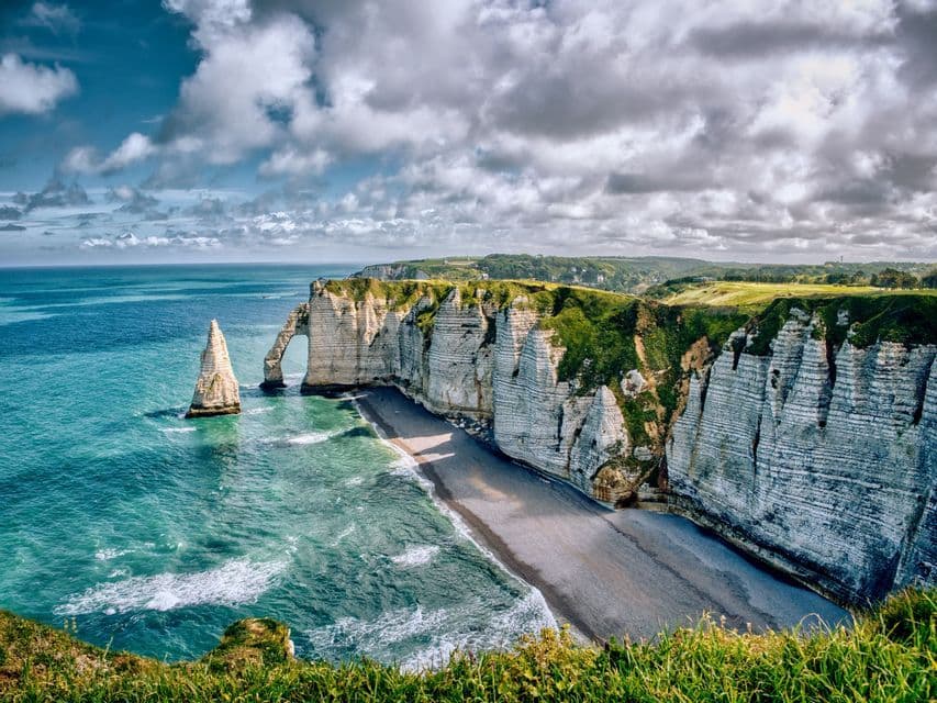 Falaises de craie blanche avec une arche naturelle et une aiguille rocheuse le long d'une côte aux eaux turquoise sous un ciel nuageux.