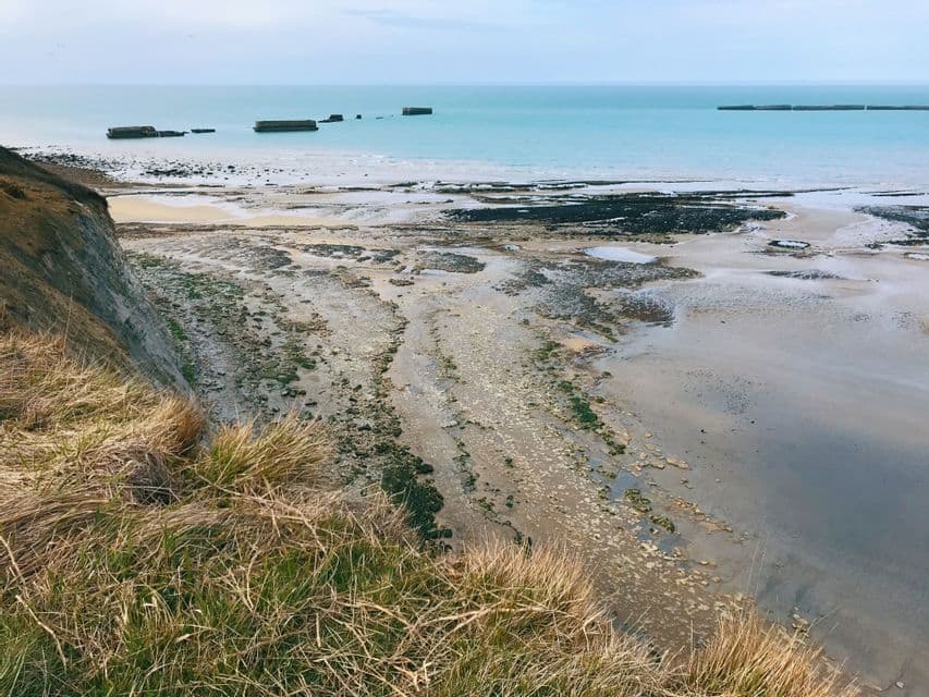 Una vista desde un acantilado cubierto de hierba revela una playa rocosa con ruinas de hormigón en el tranquilo mar turquesa durante la marea baja.