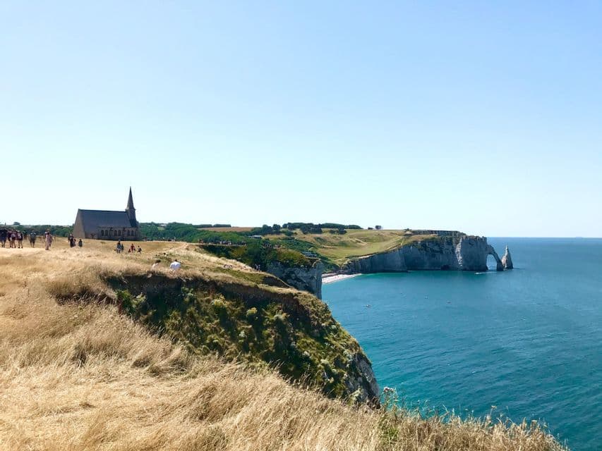 Una chiesa sorge su una scogliera erbosa che si affaccia sul mare blu, con una formazione rocciosa ad arco naturale in lontananza.