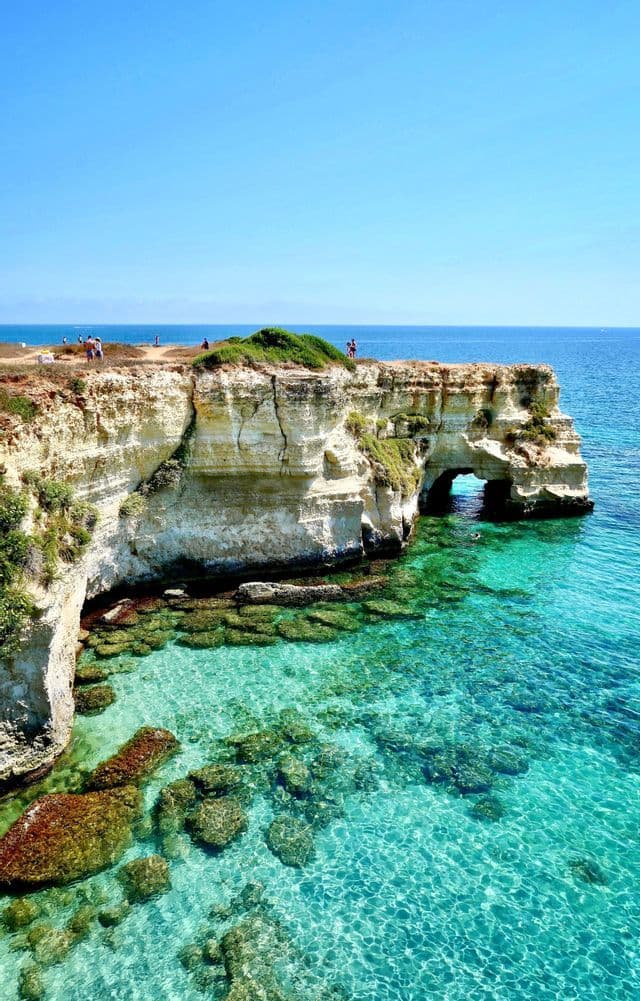 A coastal view of white cliffs and a natural rock arch over clear turquoise water on a sunny day.
