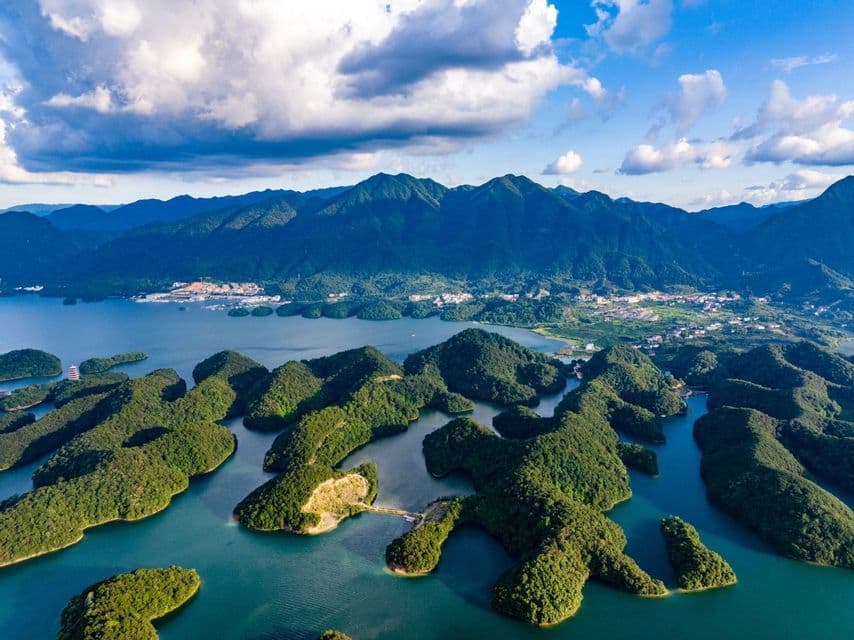An aerial view of a vast lake with numerous small, forested islands, backed by a mountain range under a partly cloudy sky.