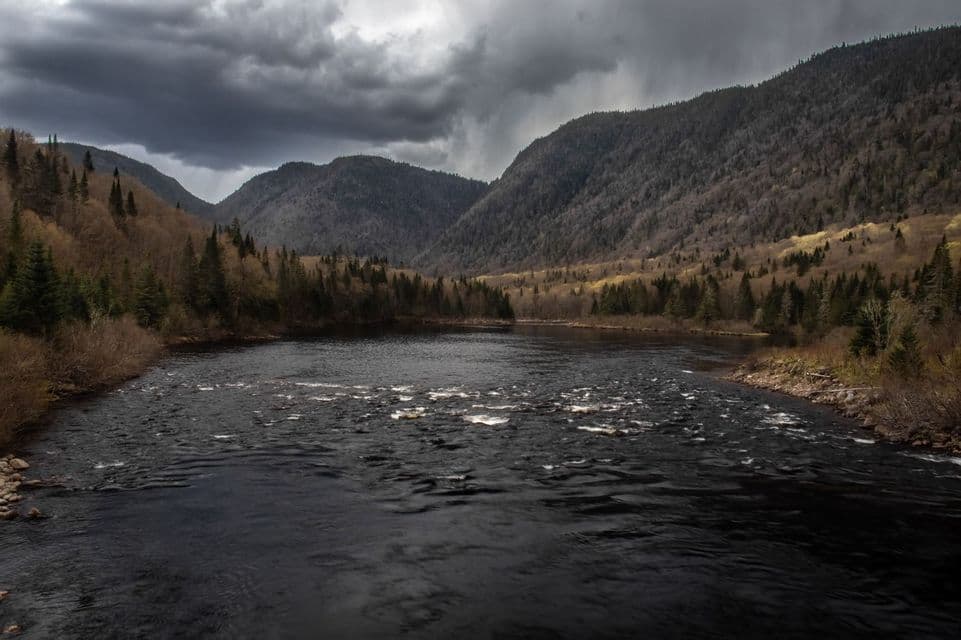 Un río oscuro fluye por un valle entre montañas boscosas bajo un cielo lleno de nubes pesadas y tormentosas.