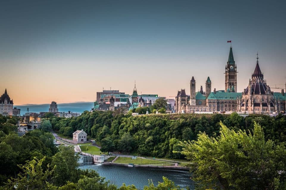 Un panorama urbain avec des bâtiments du parlement de style gothique, sur une colline boisée surplombant un réseau de canaux au coucher du soleil.
