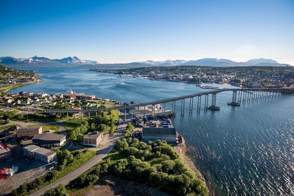 Vue aérienne d'un long pont traversant un large chenal d'eau vers une ville côtière, avec des montagnes enneigées au loin.