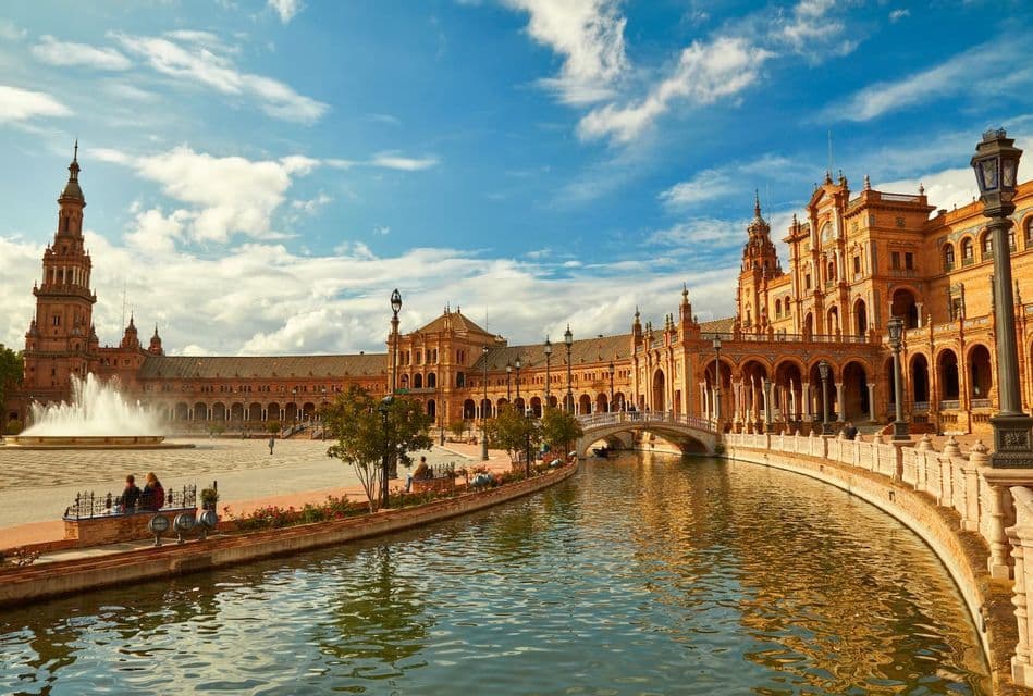 Un grand bâtiment orné avec une tour et des arches borde un canal sinueux sur une place avec une fontaine sous un ciel bleu.