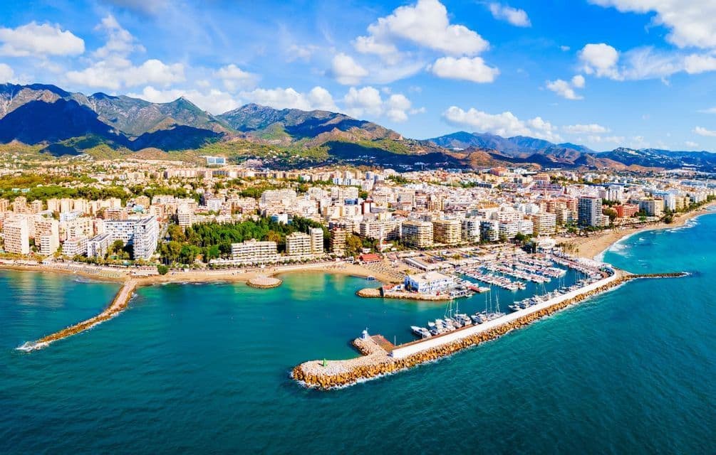 Vue aérienne d'une ville côtière avec un grand port de plaisance, une plage de sable et des montagnes en arrière-plan sous un ciel bleu nuageux.