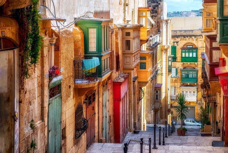 Una calle estrecha y empinada con escalones de piedra desciende entre edificios con balcones cerrados y coloridos.