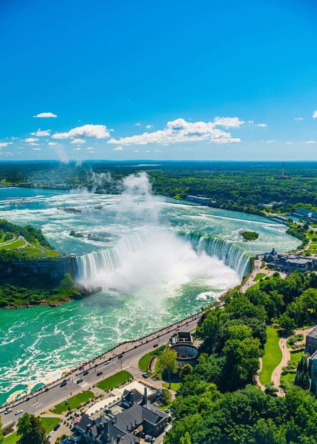 Vue aérienne d'une grande cascade en fer à cheval se jetant dans une rivière turquoise, avec une route et une ville nichées dans un paysage verdoyant luxuriant.