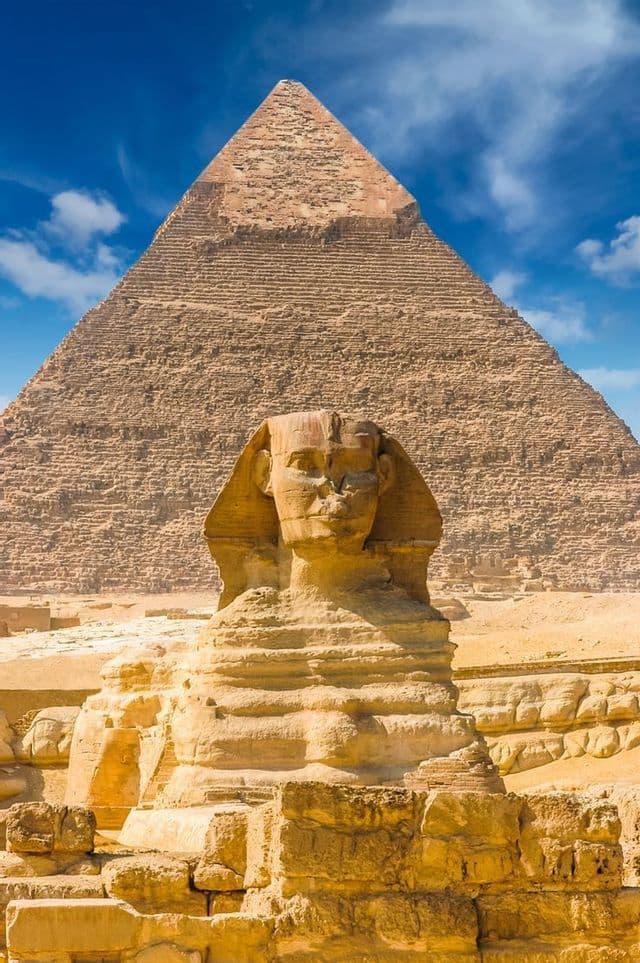 La Grande Sfinge si trova di fronte a una grande piramide nel deserto sotto un cielo azzurro con alcune nuvole.