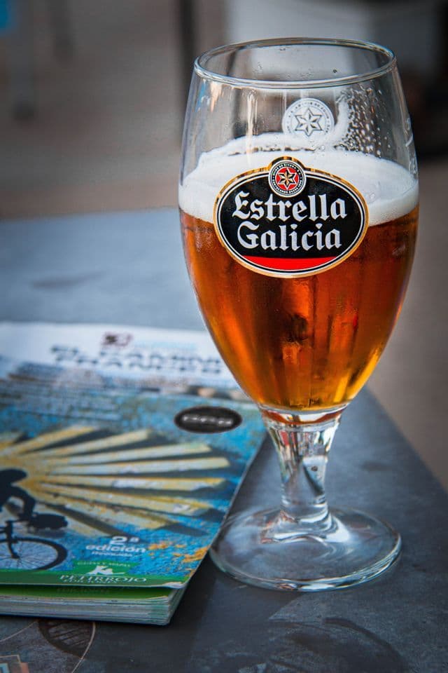 Nahaufnahme eines Glases Estrella Galicia Bier mit Schaumkrone, auf einem dunklen Tisch neben einer Zeitschrift stehend.