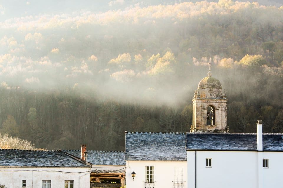 Un campanile in pietra si innalza sopra le case bianche del villaggio, con tetti in ardesia, su un fianco di collina nebbioso e boscoso.