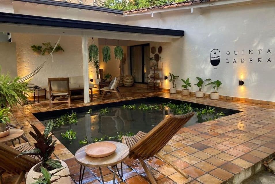 Une cour-patio carrelée avec un bassin central, des chaises longues en bois, un hamac et de nombreuses plantes en pot sous un éclairage chaleureux.