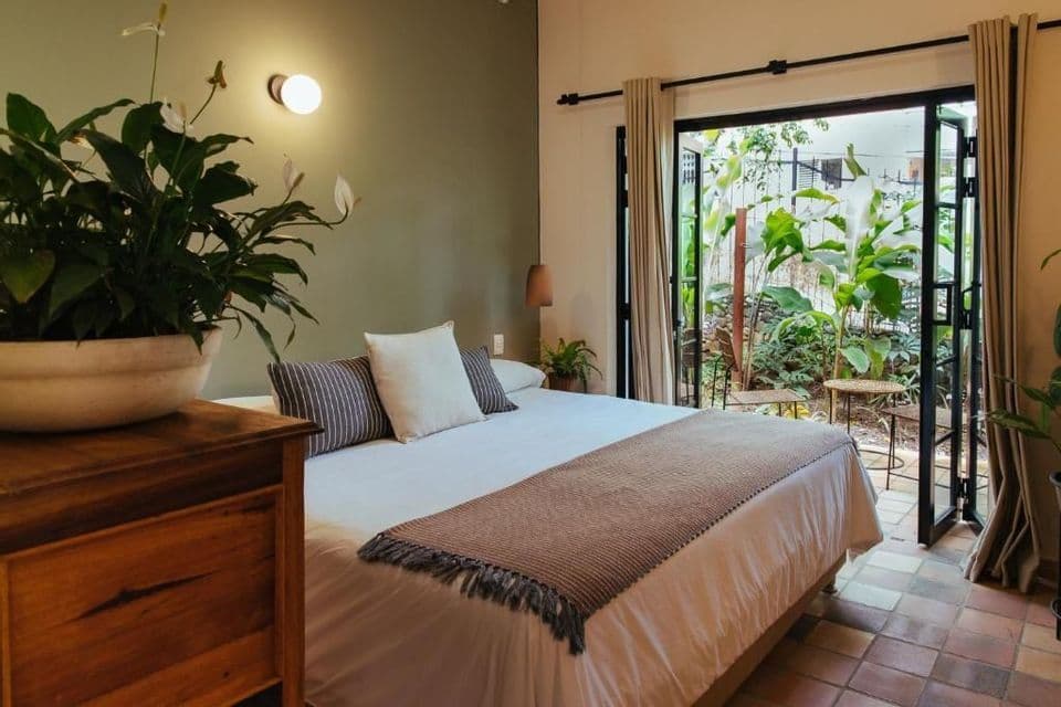 Intérieur d'une chambre avec un lit double, une grande plante en pot et des portes ouvertes donnant sur un patio verdoyant.