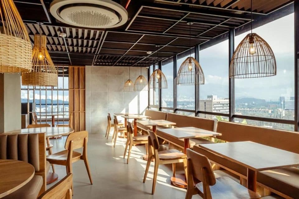 L'intérieur ensoleillé d'un restaurant moderne avec des meubles en bois, des suspensions en osier et de grandes fenêtres donnant sur un paysage urbain.