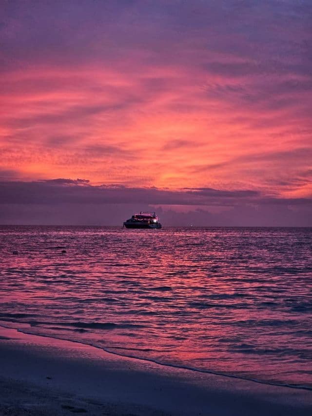 Un barco con luces flota en el mar bajo un cielo rosado y púrpura al atardecer, con una costa en primer plano.