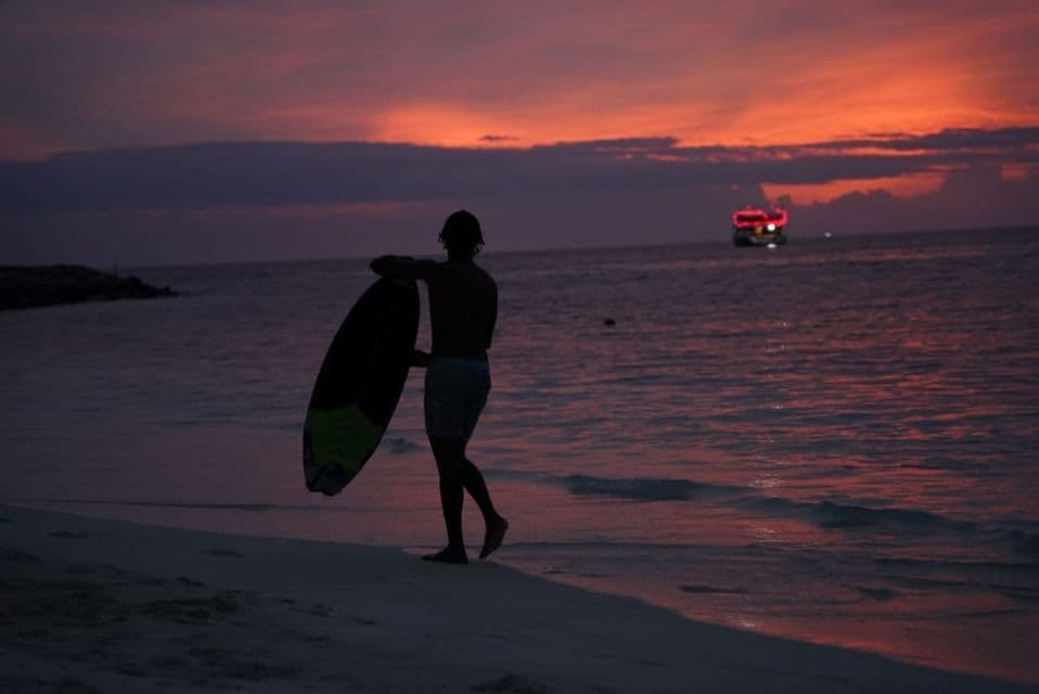 Una persona silueteada con una tabla de surf camina por la orilla al atardecer, con un bote en el agua a lo lejos.