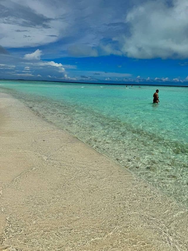Un hombre de pie en agua poco profunda, clara y turquesa, con una playa de arena en primer plano bajo un cielo nublado.