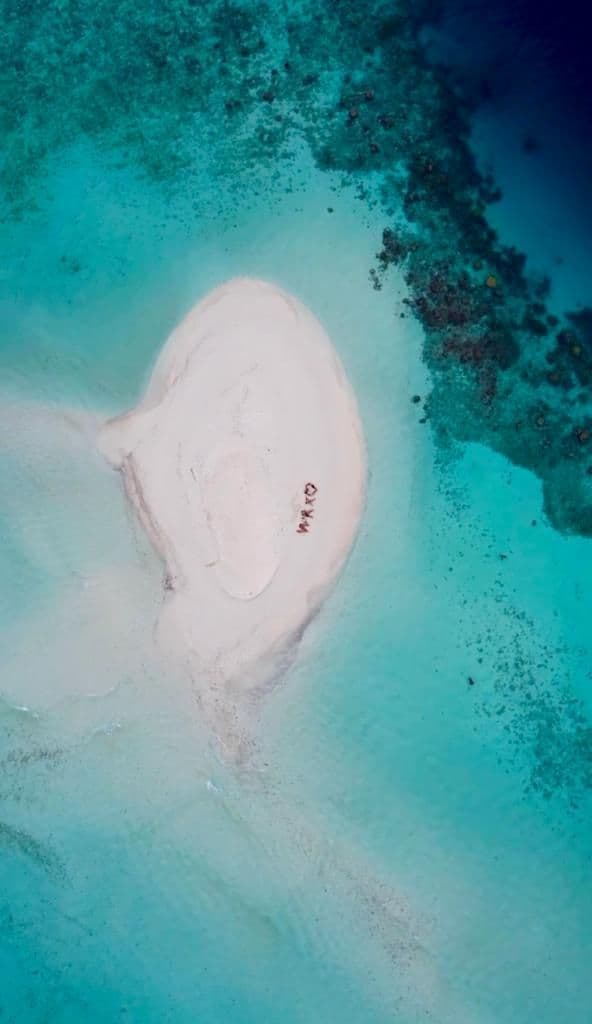 Vista aerea di un gruppo WeRoad in viaggio che forma lettere con i corpi su una lingua di sabbia bianca in acqua turchese.