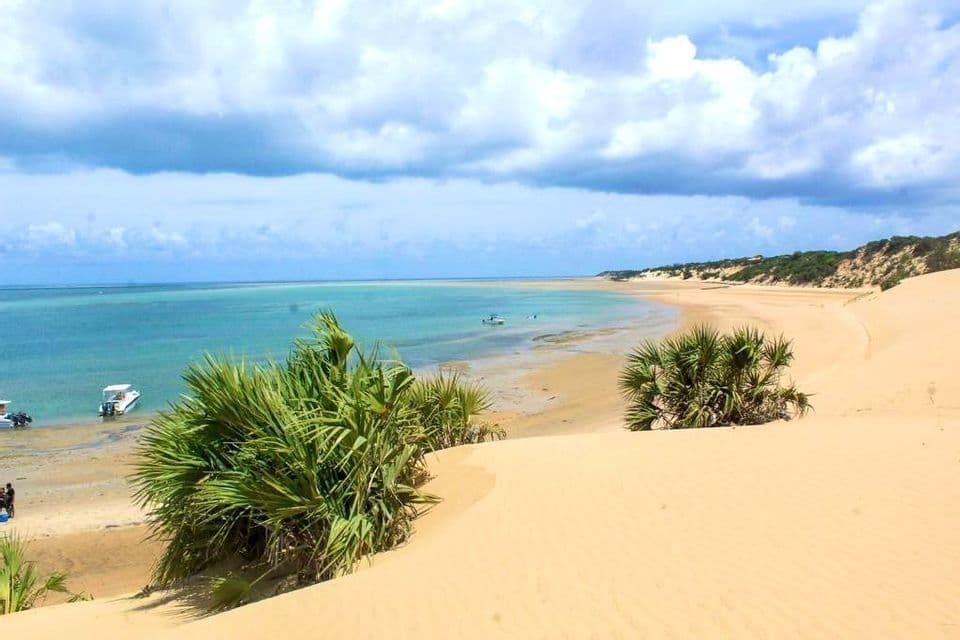 Blick von einer Sanddüne mit grünen Büschen auf eine türkisfarbene Bucht mit am Strand ankernden Booten unter bewölktem Himmel.