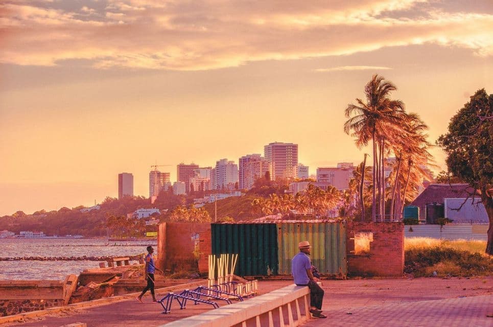 Un uomo siede su un muro a guardare il tramonto su uno skyline di città costiera con palme e il mare in primo piano.