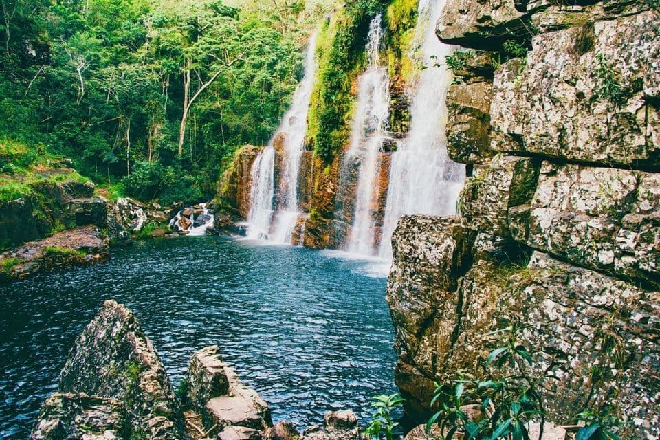 Una cascata si getta da una scogliera rocciosa terrazzata e muschiosa in una pozza blu profonda, circondata da una lussureggiante foresta verde.